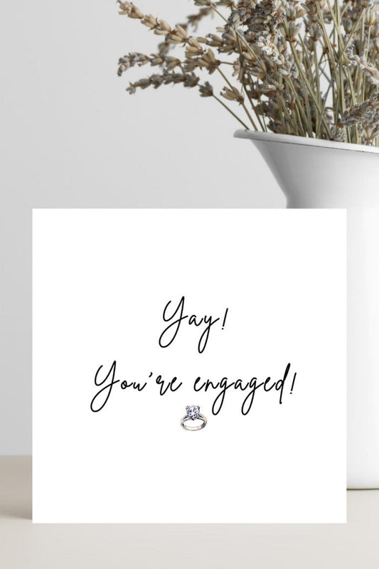 A white square greeting card with the words 'Yay! You're engaged!' in black script and a small ring illustration, styled on a tabletop next to a white vase with dried flowers.