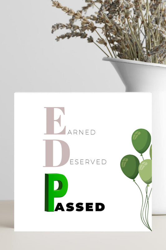 Card with 'Earned, Deserved, Passed' text and green balloons on a white surface with a plant in the background.