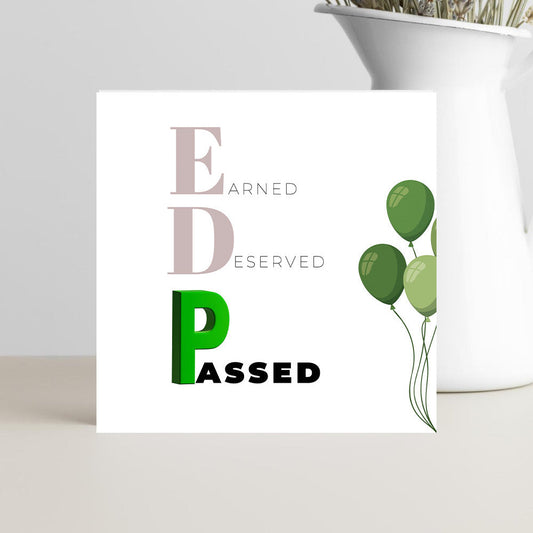 Card with 'Earned, Deserved, Passed' text and green balloons on a white surface with a plant in the background.