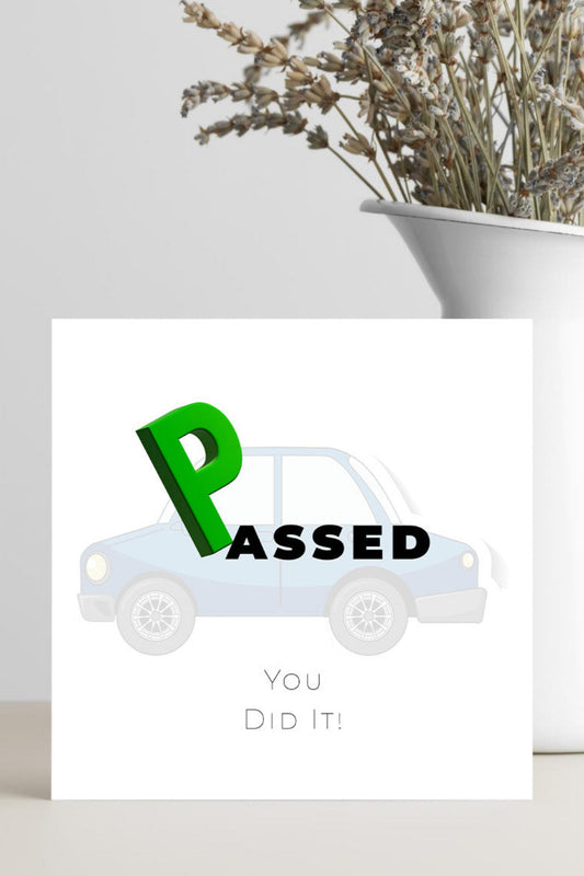 A square white greeting card standing on a light surface, printed with a light blue car, a large green 'P', and the words 'PASSED' and 'You Did It!'. A white vase with dried stems sits behind the card against a light gray wall.