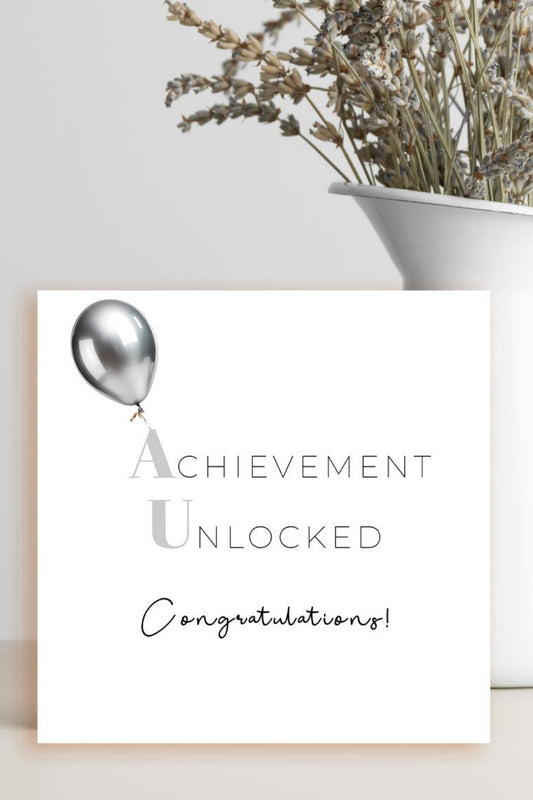 A square white congratulations card with a silver balloon graphic and the words 'Achievement Unlocked' and 'Congratulations!' in gray and black script, styled in front of a white vase with dried flowers against a light gray background.