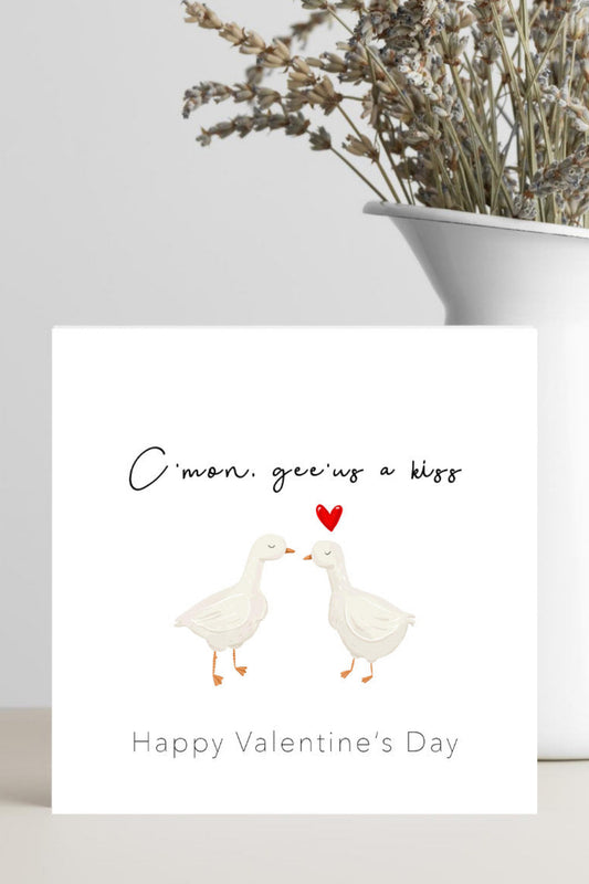 A square Valentine's Day greeting card with a white background, featuring an illustration of two white ducks facing each other with a small red heart between them. The text above reads 'C'mon give us a kiss' and below reads 'Happy Valentine's Day'.