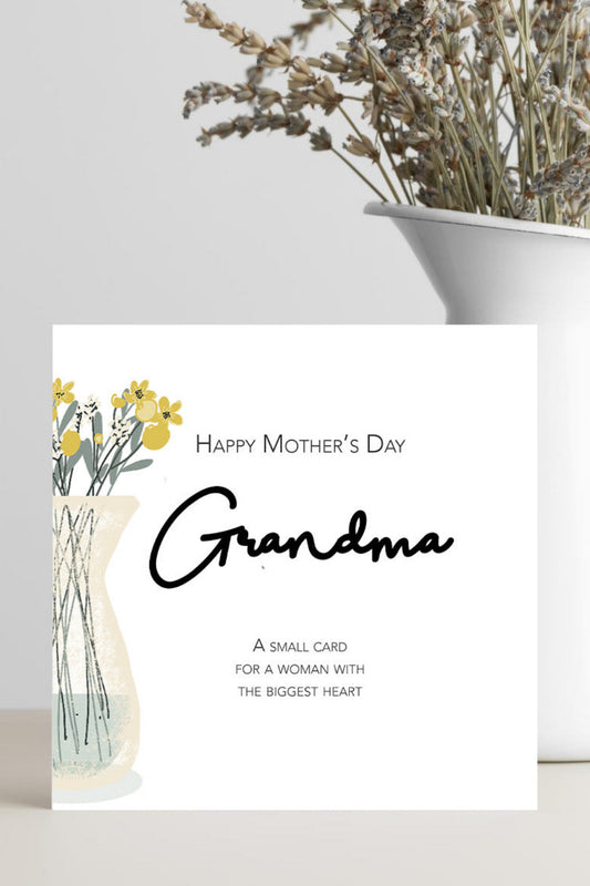 A square Mother's Day card addressed to Grandma, featuring a floral illustration of yellow flowers in a vase and elegant black script text, styled on a light surface next to a white ceramic pitcher filled with dried flowers.