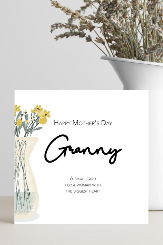A square white greeting card with 'Happy Mother's Day Granny' printed in black script and serif text, plus a small floral illustration, standing on a light surface beside a white vase with dried flowers.