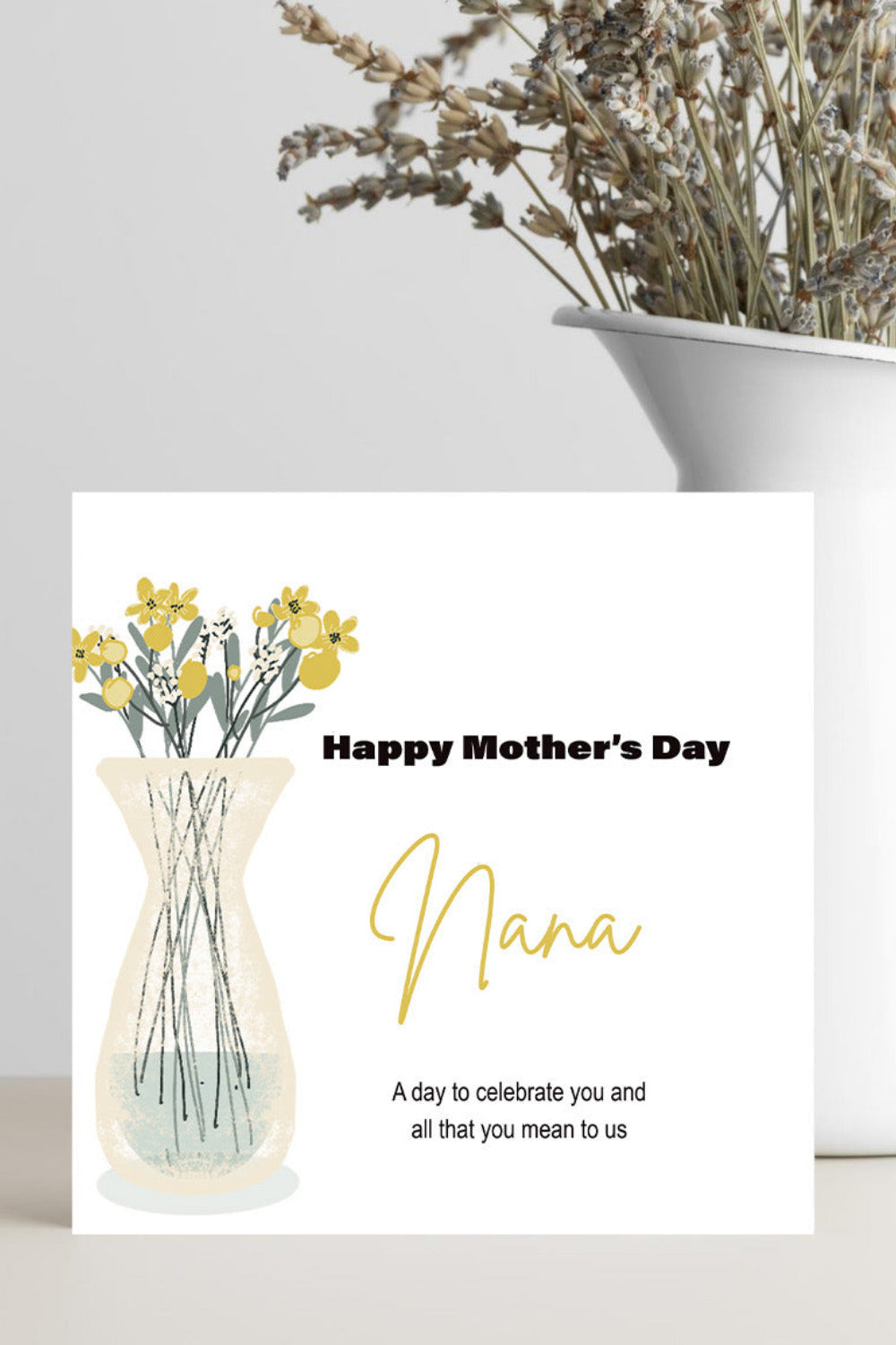 A Mother's Day greeting card with a watercolor-style vase of yellow flowers and the text 'Happy Mother's Day Nana' stands on a light surface. A white ceramic vase with dried flowers sits behind it against a light gray wall.