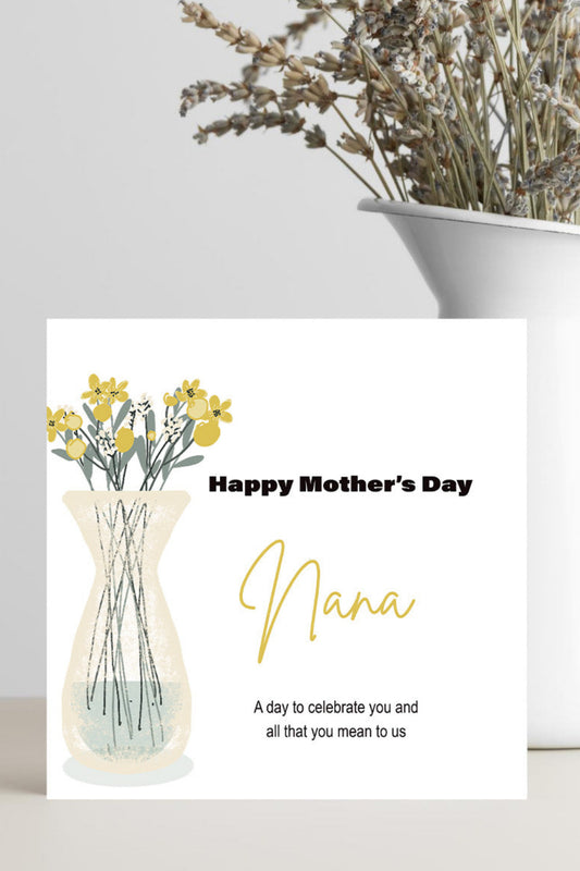 A Mother's Day greeting card with a watercolor-style vase of yellow flowers and the text 'Happy Mother's Day Nana' stands on a light surface. A white ceramic vase with dried flowers sits behind it against a light gray wall.