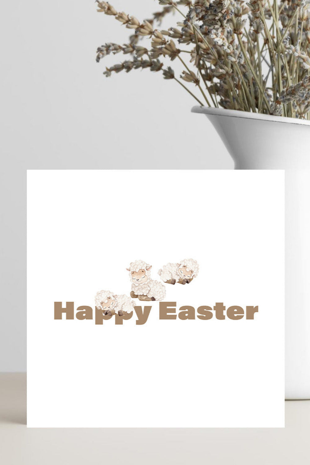 A white square greeting card with a brown 'Happy Easter' text and small sheep illustration, standing on a tabletop beside a white vase with dried flowers against a light gray wall.