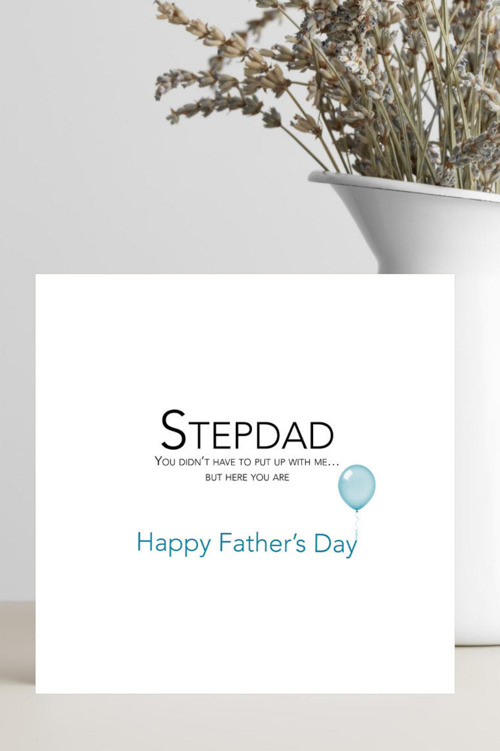 A white greeting card with the text 'STEPDAD' and 'Happy Father's Day' in blue, styled on a light surface next to a white vase with dried flowers.