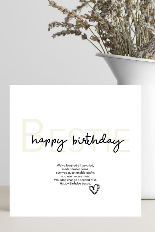 A white square greeting card with the words 'happy birthday' in black script and small printed text below, styled on a light surface beside a white vase with dried flowers.