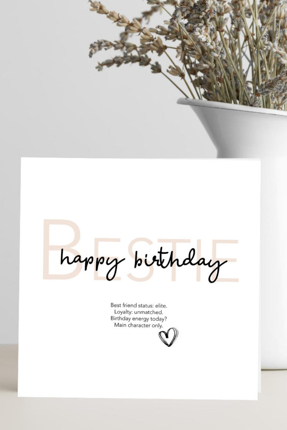 Greeting card with 'Bestie, happy birthday' text on a white surface with a vase of dried flowers.