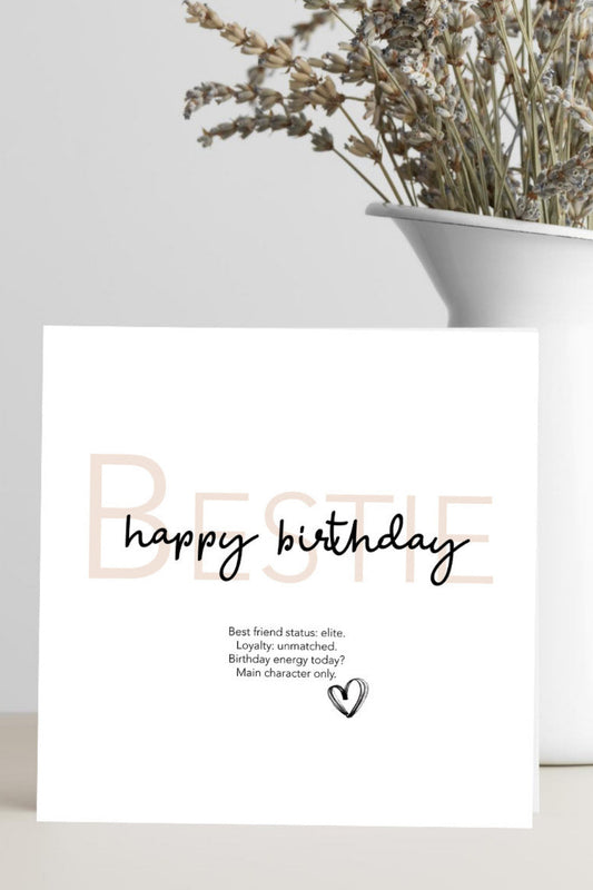 Greeting card with 'Bestie, happy birthday' text on a white surface with a vase of dried flowers.