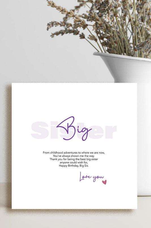 A square white birthday card with purple and black text reading 'Big Sister' and a short message, styled on a tabletop beside a white vase with dried flowers against a light gray wall.