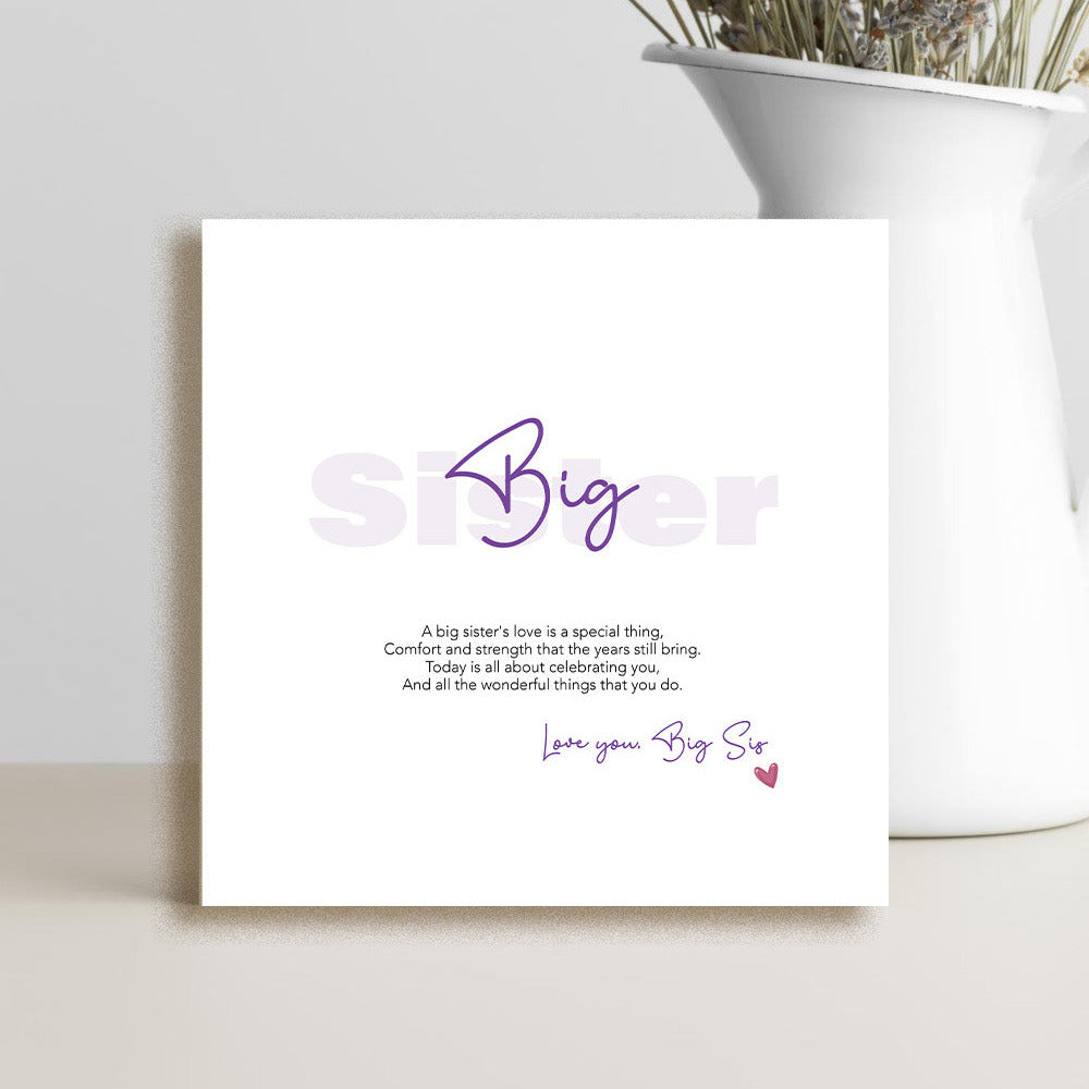 A square white greeting card with purple and black text reading 'Big Sister' and a short message, styled leaning against a light wall beside a white vase with dried flowers.