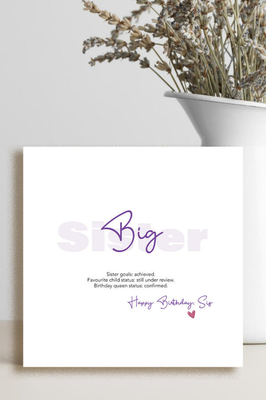 A square white birthday card with purple and gray text reading 'Big Sister' and 'Happy Birthday Sis,' styled on a light surface beside a white vase with dried flowers.