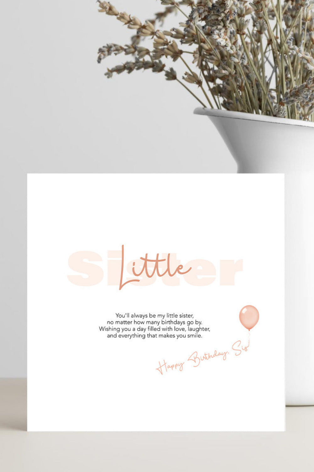 A square white birthday card with peach-colored text reading 'Little Sister' and a short birthday message, styled on a tabletop next to a white ceramic vase with dried flowers.