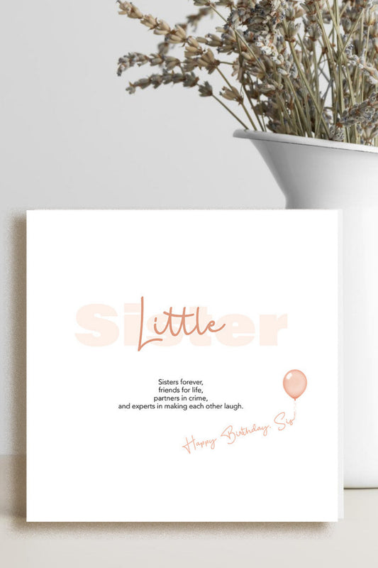 A square birthday card with a white background and peach-colored text reading 'Little Sister' and 'Happy Birthday!' with a small balloon illustration, styled on a tabletop next to a white vase with dried flowers.