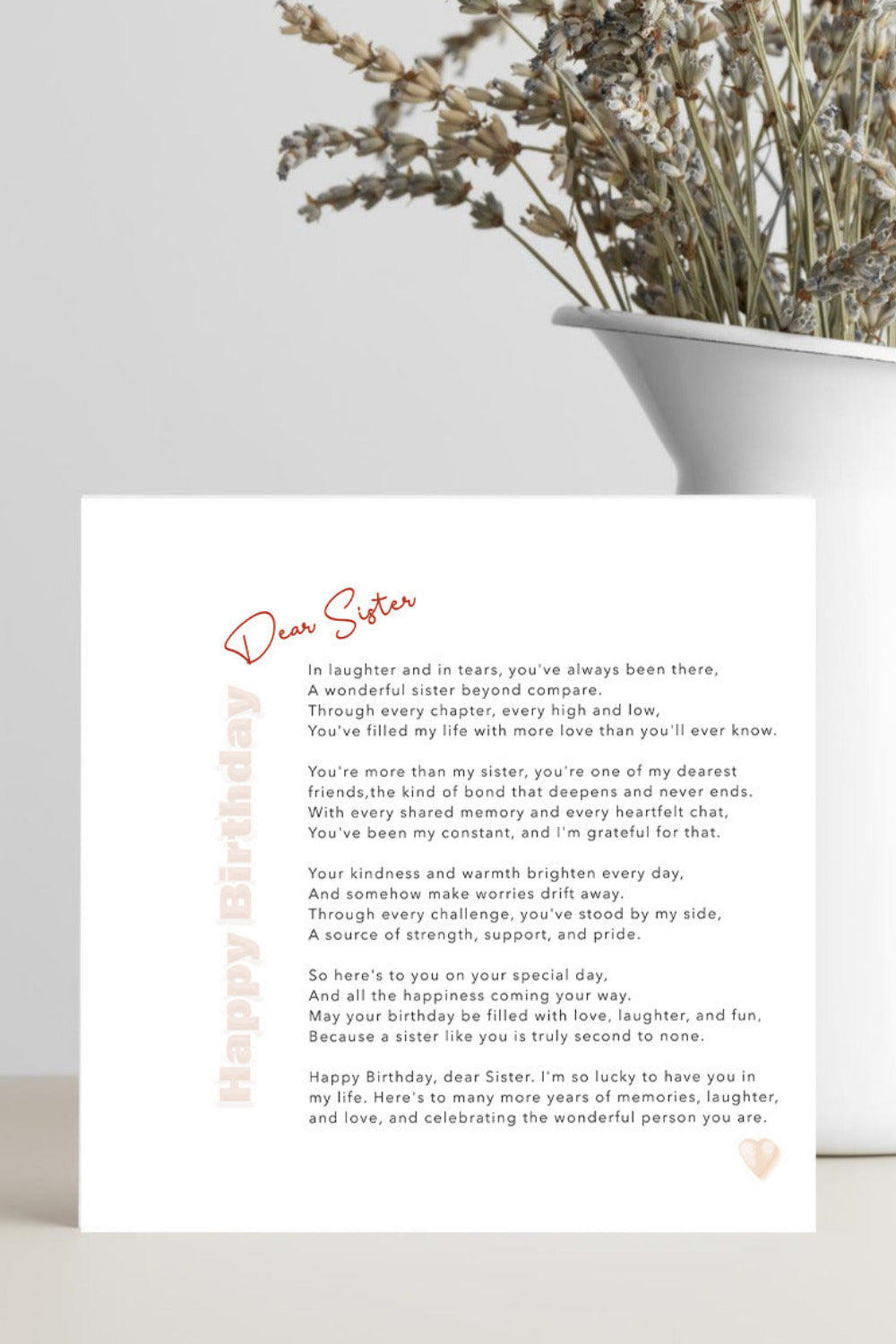 A square white birthday card with a poem titled 'Dear Sister' and 'Happy Birthday' in red script, placed on a light surface next to a white vase containing dried flowers.