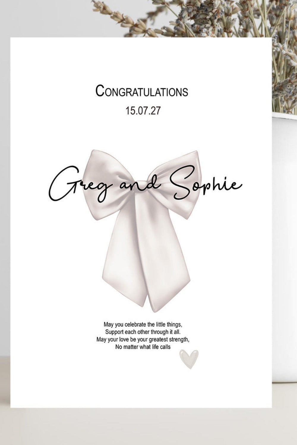 A personalized congratulations card with a white background, featuring a large bow illustration and the names 'Greg and Sophie' in black script. The card is styled on a light surface next to a white vase with dried flowers.