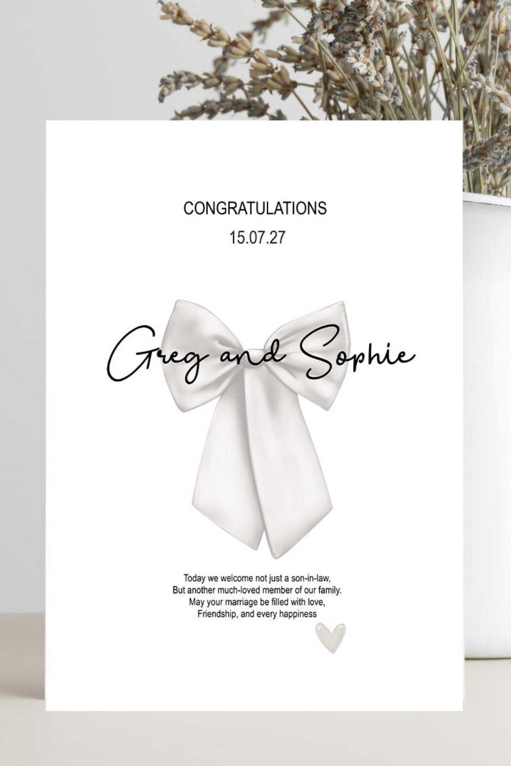 A white congratulations card with a large white bow illustration and the names 'Greg and Sophie' in black script, photographed leaning against a light gray background with dried flowers in a white vase.
