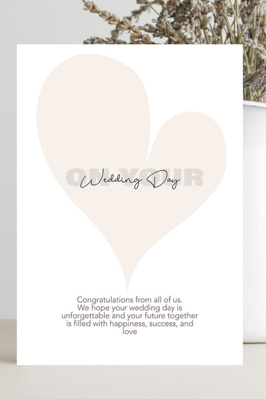 A white wedding anniversary greeting card with a large pale blush heart, the words 'Wedding Day' across the center, and a short message of congratulations at the bottom, styled on a light surface beside dried flowers in a white vase.