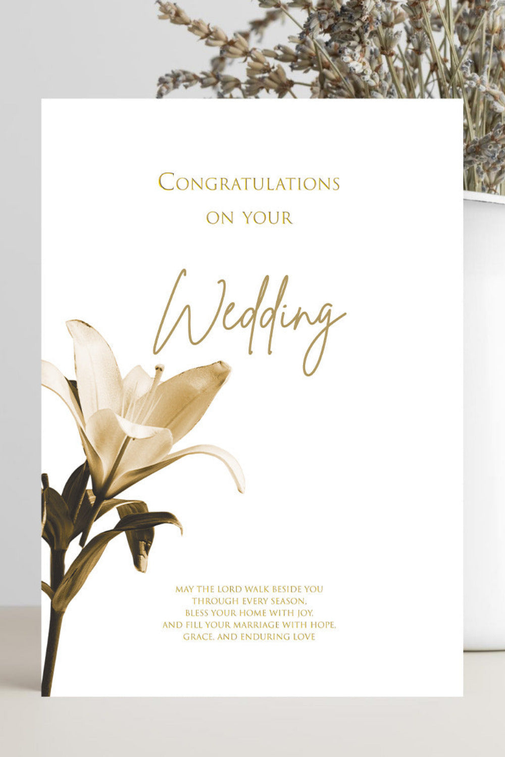 A white wedding congratulations card with elegant gold typography and a gold-toned lily illustration, styled in a flat-lay setting next to a white vase with dried flowers.