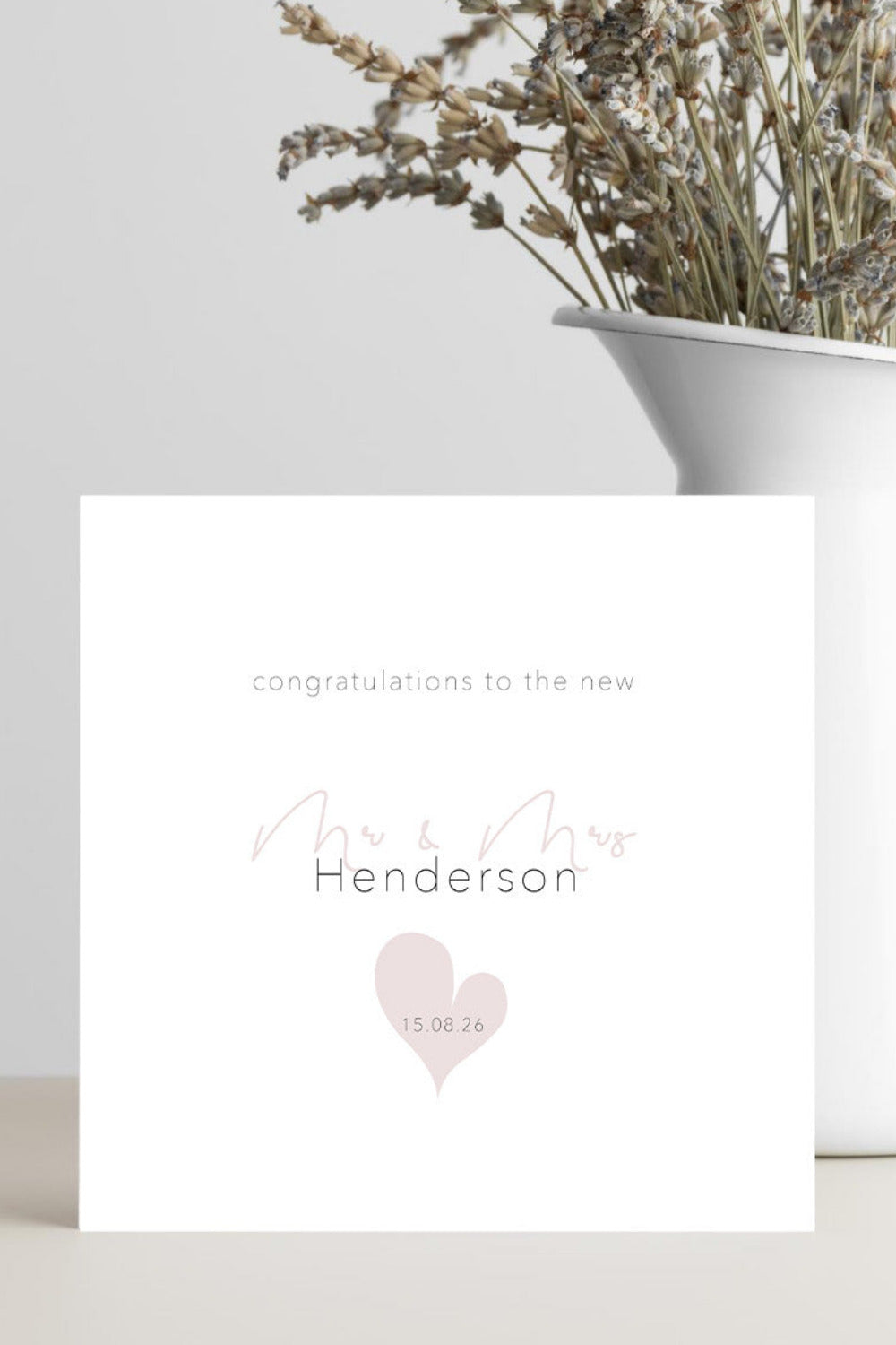 A square white congratulations card with elegant script and serif text reading 'congratulations to the new Mr & Mrs Henderson' and a pale pink heart with the date '15.08.