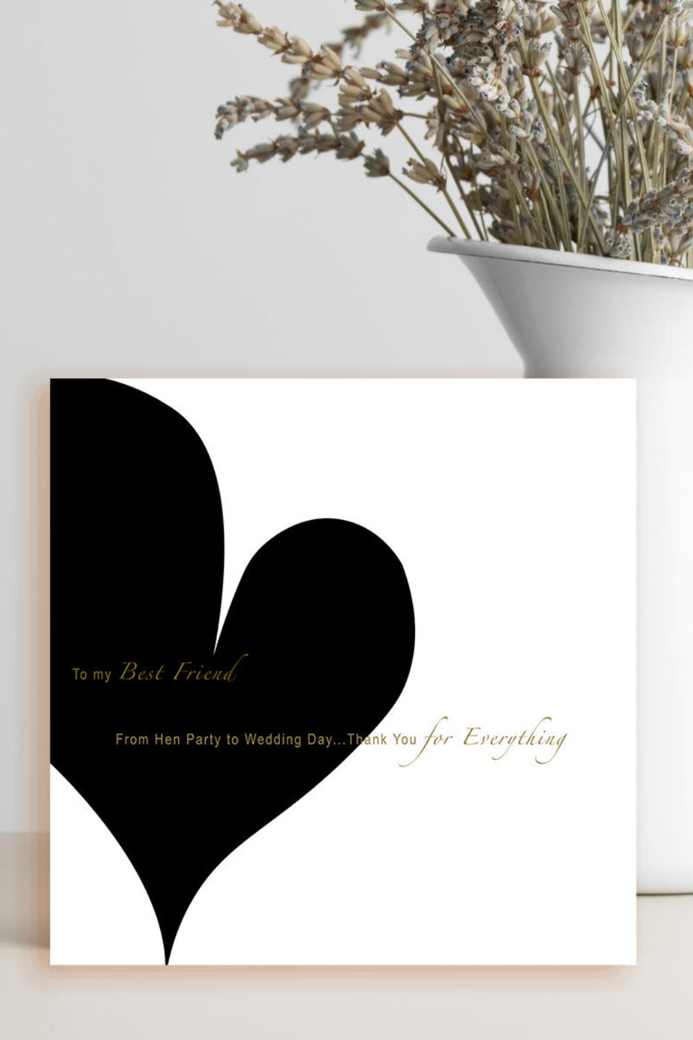 A square white greeting card with a large black heart and gold script text, styled on a light surface next to a white vase with dried flowers.