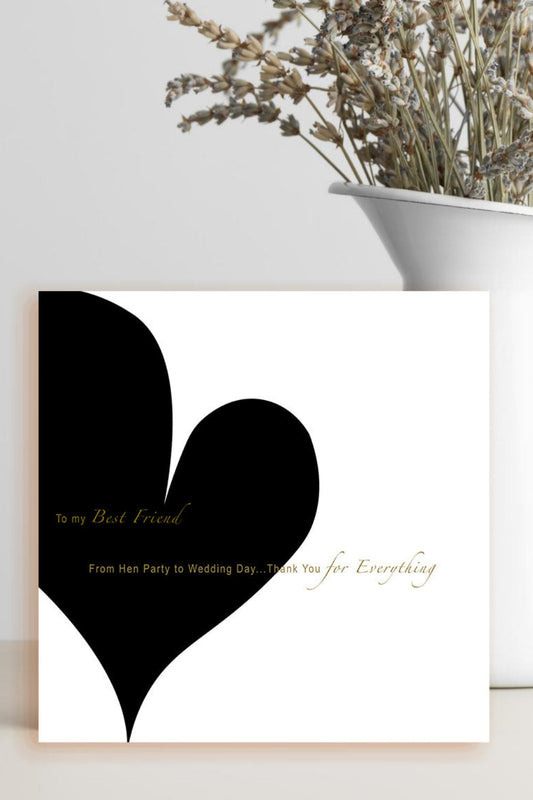 A square white greeting card with a large black heart and gold script text, styled on a light surface next to a white vase with dried flowers.