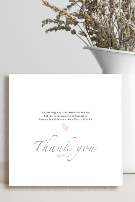 A square white wedding thank you card with elegant gray script and a small pink heart, styled on a tabletop next to a white vase with dried flowers.
