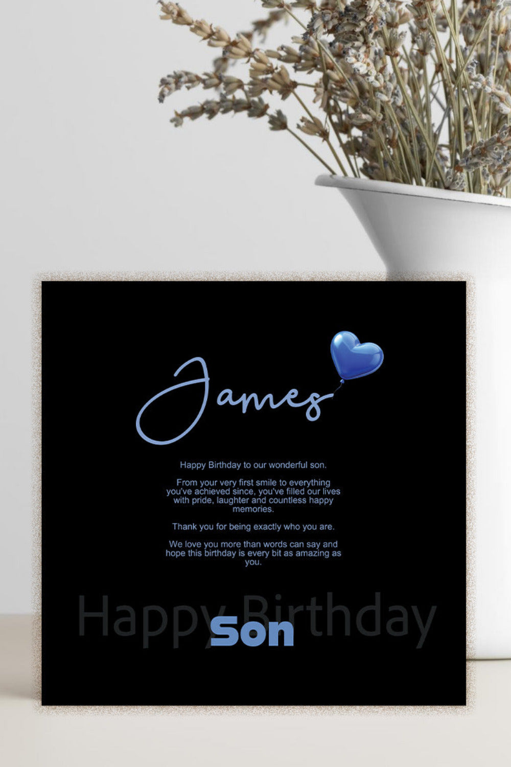 A black personalized birthday card with blue text and a blue heart, styled on a light surface next to a white vase with dried flowers.