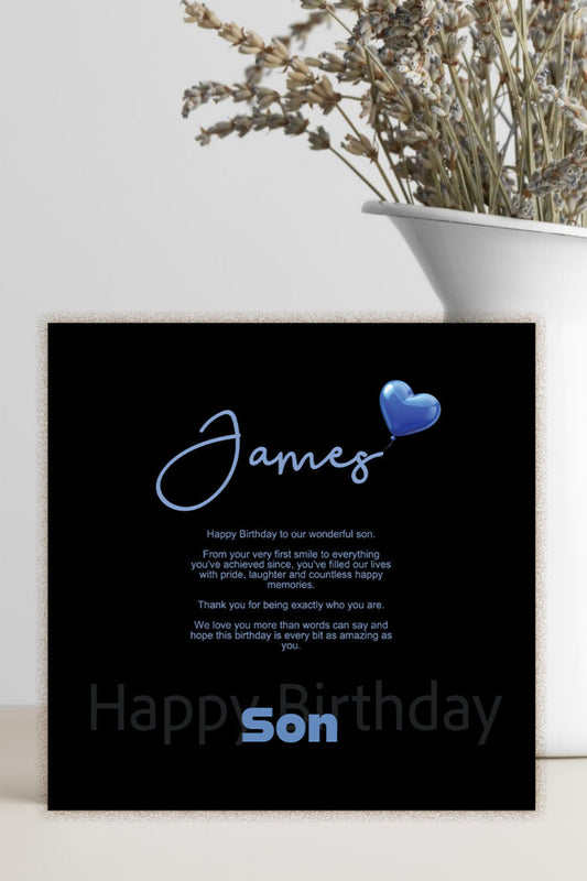 A black personalized birthday card with blue text and a blue heart, styled on a light surface next to a white vase with dried flowers.
