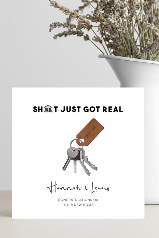 A square white greeting card standing on a light surface, printed with the phrase 'SHIT JUST GOT REAL' above an illustration of keys with a brown tag, and personalized names 'Hannah & Lewis' with the message 'CONGRATULATIONS ON YOUR NEW HOME' below. A white vase with dried flowers sits behind the card against a light gray wall.