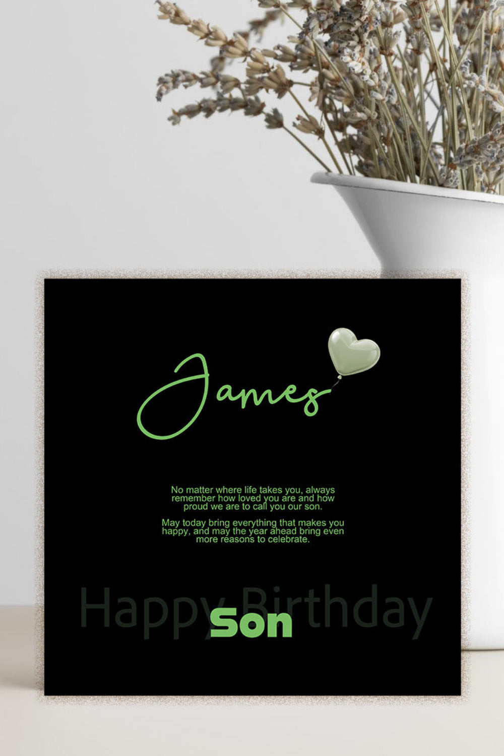 A personalized black square card with green text reading 'James' and 'Happy Birthday Son' stands on a light surface beside a white vase with dried flowers.