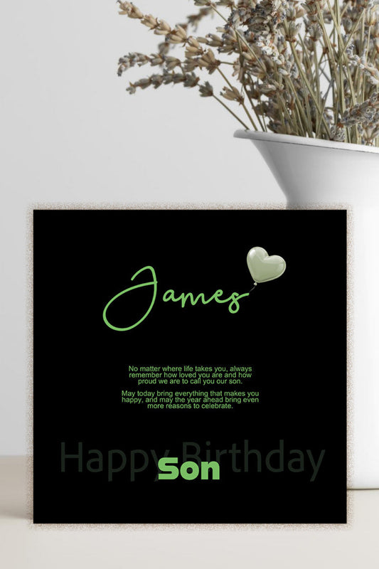 A personalized black square card with green text reading 'James' and 'Happy Birthday Son' stands on a light surface beside a white vase with dried flowers.