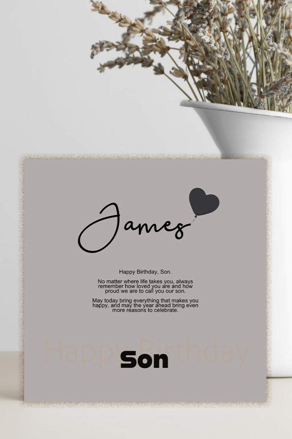 A personalized birthday card with a light gray background and black text, styled on a light beige surface next to a white ceramic vase with dried flowers.