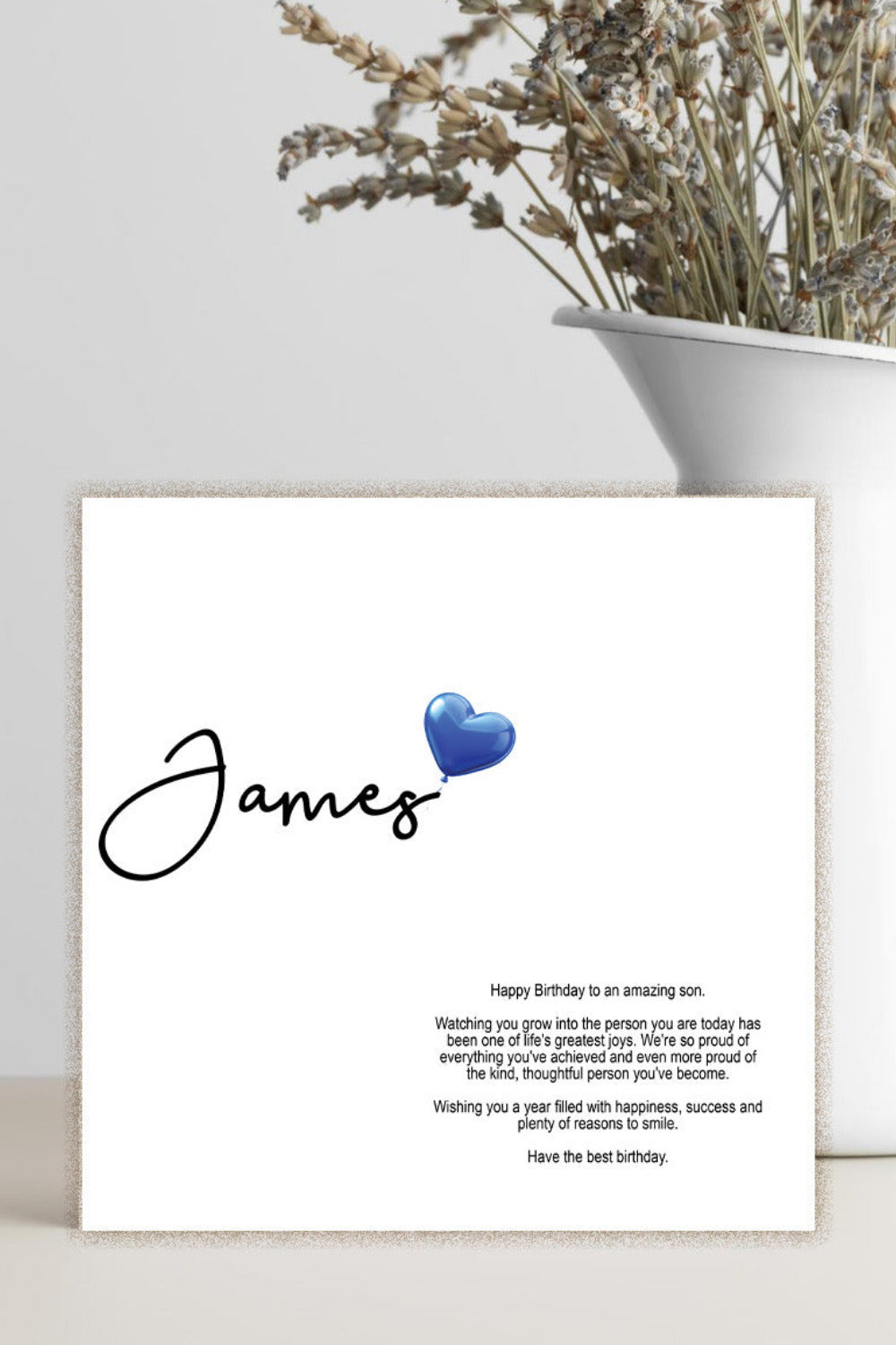 A personalized birthday card with the name 'James' and a blue heart, displayed in a white ceramic vase with dried flowers against a light gray background.