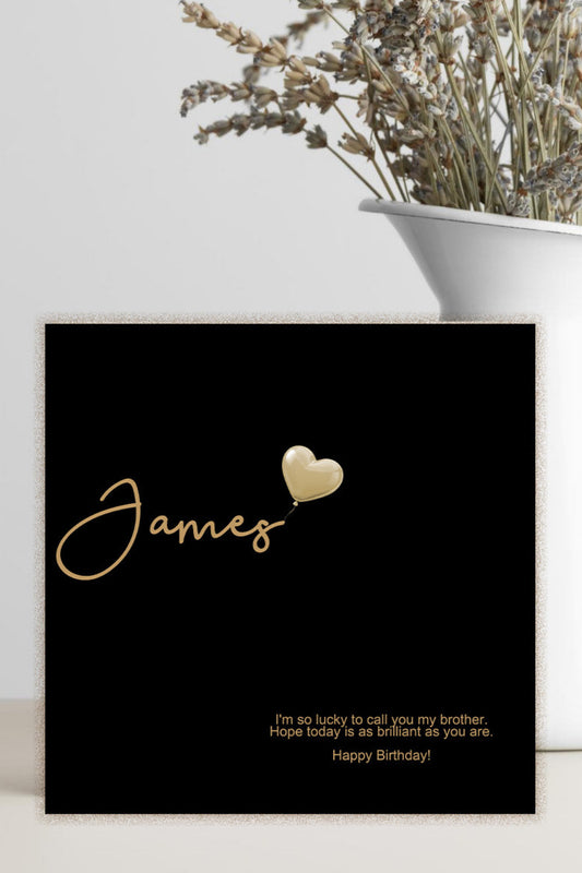 A black personalized birthday card with gold foil script reading 'James' and a small gold heart, styled on a light surface beside a white vase with dried flowers.
