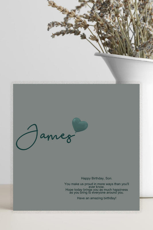 A personalized birthday card for a son, featuring a solid gray cover with the name 'James' in teal script and a small heart, displayed next to a white vase with dried flowers.