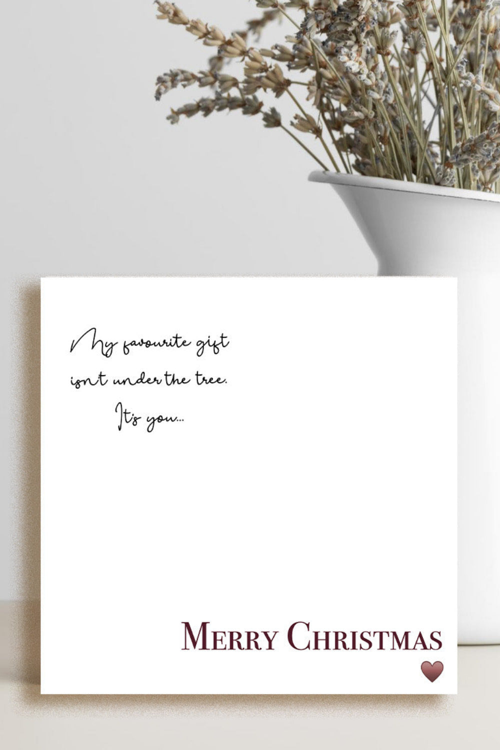 A square white Christmas greeting card with elegant black script text and 'MERRY CHRISTMAS' in burgundy at the bottom, styled on a light beige surface next to a white ceramic vase with dried flowers.