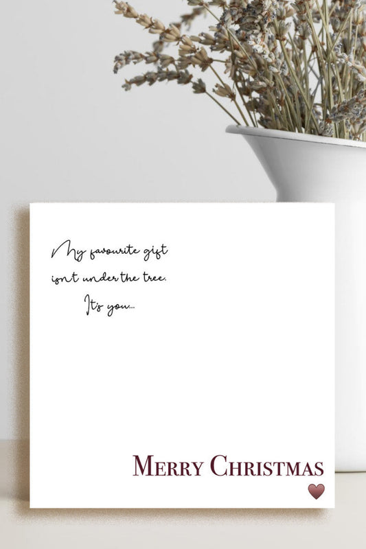 A square white Christmas greeting card with elegant black script text and 'MERRY CHRISTMAS' in burgundy at the bottom, styled on a light beige surface next to a white ceramic vase with dried flowers.
