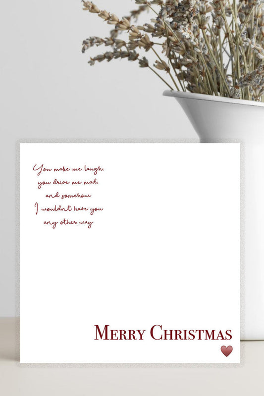 A white Christmas greeting card with red text reading 'Merry Christmas' and a small red heart, styled on a light surface beside a white vase with dried flowers against a pale gray background.