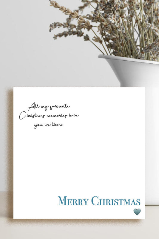 A white Christmas greeting card with black cursive text and blue 'MERRY CHRISTMAS' lettering, styled on a light surface next to a white vase with dried flowers.