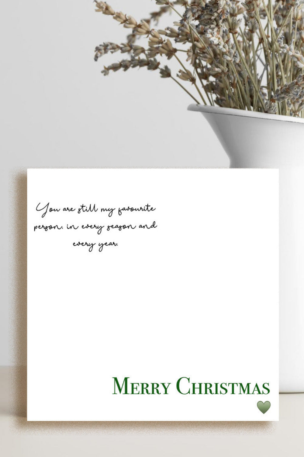 A white Christmas greeting card with black script text and green 'Merry Christmas' lettering, styled on a light beige surface next to a white ceramic vase with dried flowers.