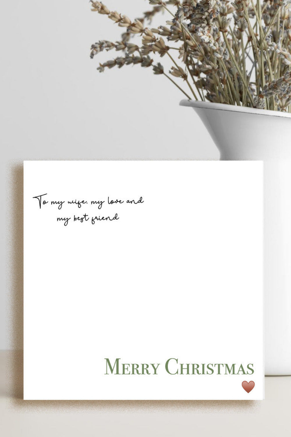 A square white Christmas greeting card with elegant black script text at the top and 'Merry Christmas' in olive green at the bottom, styled next to a white ceramic vase with dried lavender on a light beige surface.