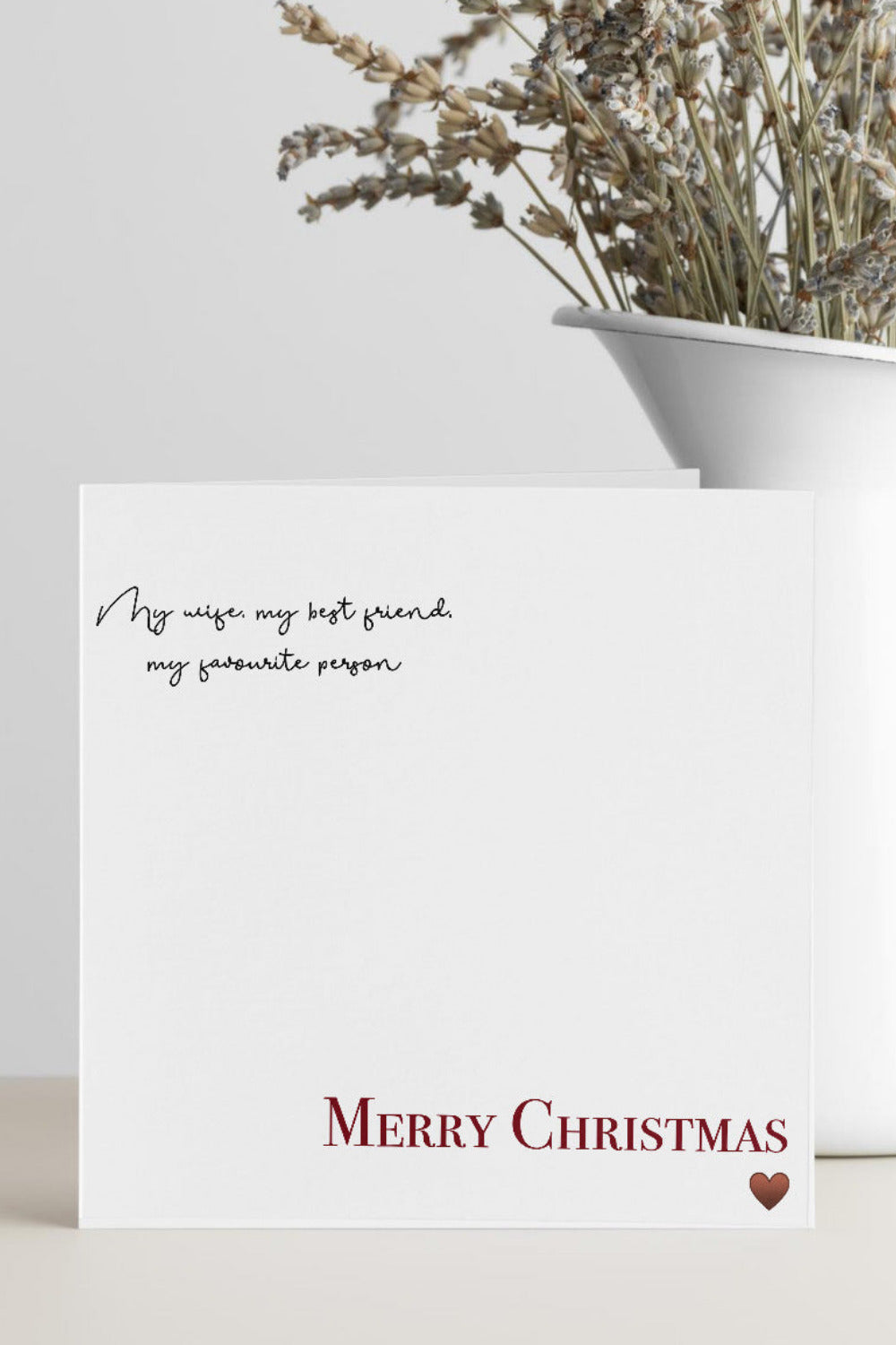 A white square greeting card with elegant black script text at the top and 'MERRY CHRISTMAS' in red serif font at the bottom, styled on a light beige surface next to a white ceramic vase with dried lavender.