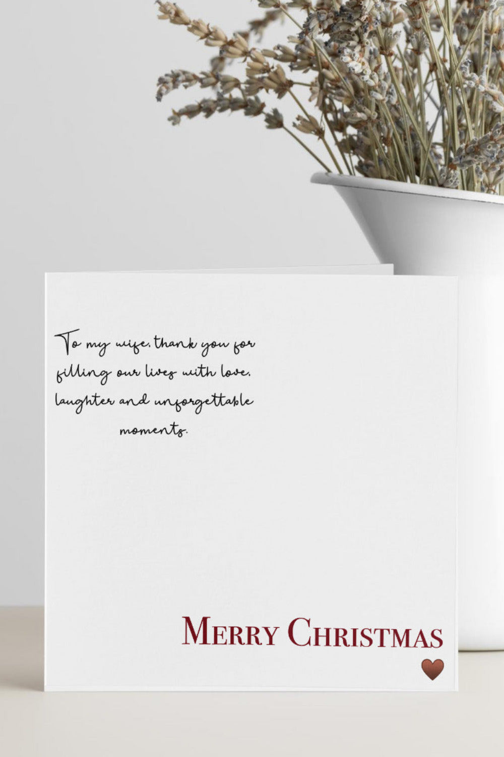 A white Christmas greeting card with a black handwritten-style message and 'MERRY CHRISTMAS' in red at the bottom, standing on a light surface beside a white vase with dried flowers.