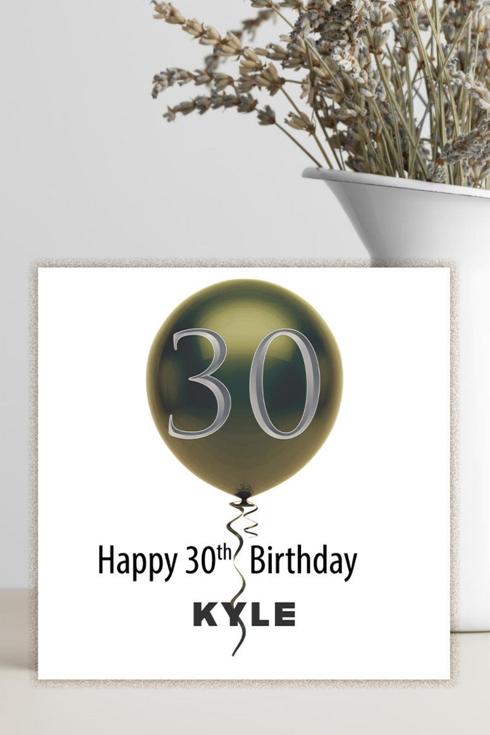 A square white birthday card with a metallic gold balloon graphic containing the number 30 and the text 'Happy 30th Birthday KYLE', displayed on a light surface next to a white vase with dried flowers.