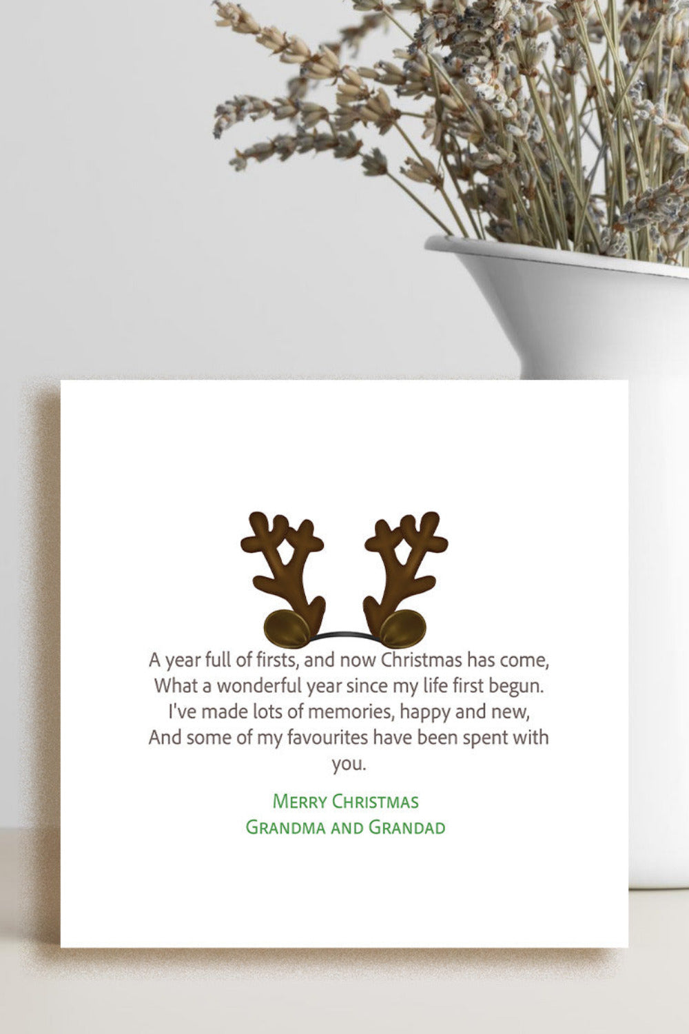 A square white Christmas greeting card with a poem and two brown reindeer antlers, styled on a light surface next to a white vase with dried flowers.