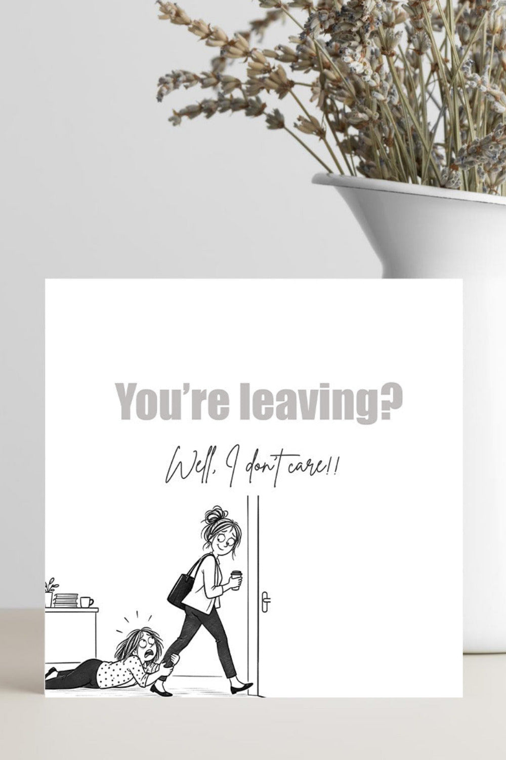 A square white greeting card with a black line drawing and the text 'You're leaving? Well, I don't care!' stands on a light surface. A white vase with dried flowers is blurred in the background.
