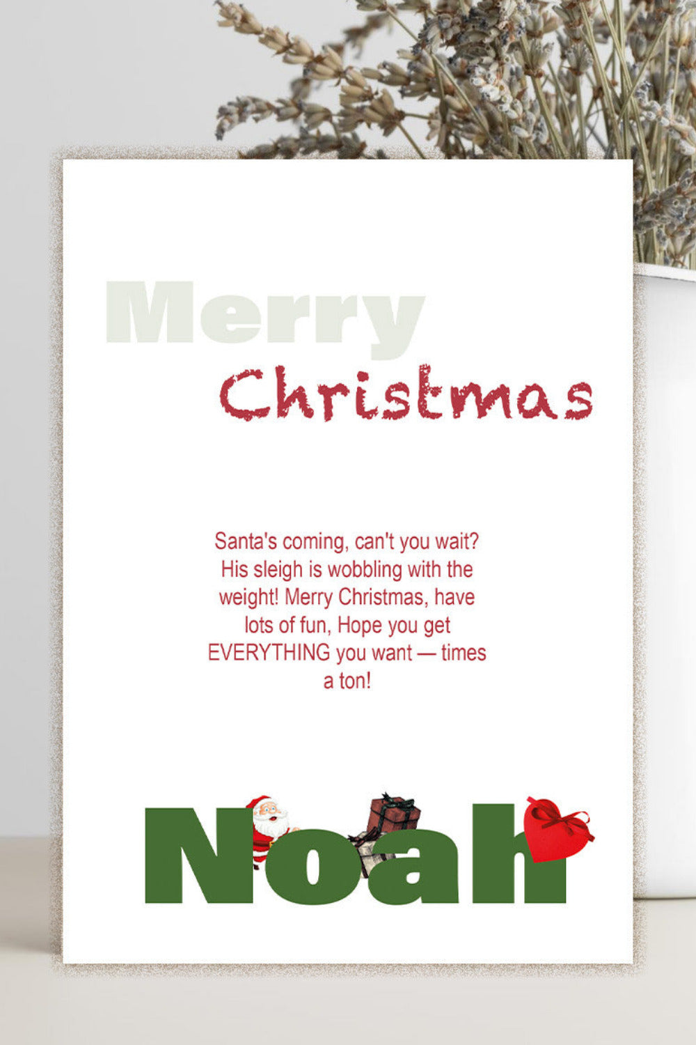 A personalized Christmas greeting card with a white background, featuring the text 'Merry Christmas' in muted green and red, a short holiday message, and the name 'Noah' printed in large green letters at the bottom with small decorative illustrations. The card is displayed standing upright on a light surface next to a white vase with dried flowers.